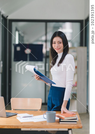 Professional millennial businesswoman in casual attire holding a clipboard with financial analysis documents, standing in the office. vertical 102391395
