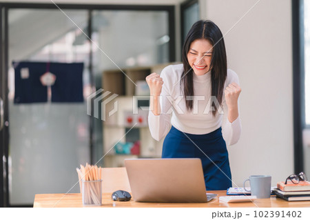 Happy and excited, a young asian woman student or office worker, celebrates achieving her goal of winning an online competition or receiving good news via email by raising hands in euphoria. 102391402