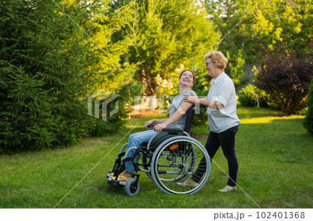 An elderly woman is carrying an adult daughter sitting in a wheelchair. Walk outdoors.  102401368