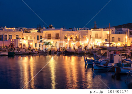 Traditional Greek fishing village and harbour after sunset, boats moored by jetty, street lamps and whitewashed houses alongside. 102403174