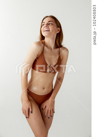 Portrait with young, smiling girl, woman in swimsuit using sunscreen cosmetic, spf on belly over white color studio background Portrait with young, smiling girl, woman in swimsuit using sunscreen cosmetic, spf on belly over white color studio background 102408803