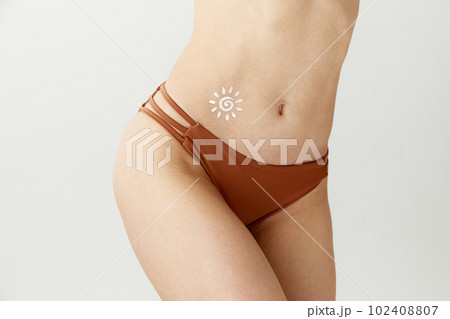 Cropped image of slim female body with light skin in swimsuit with sunscreen on belly over white color studio background. Sun protection lotion, cream 102408807