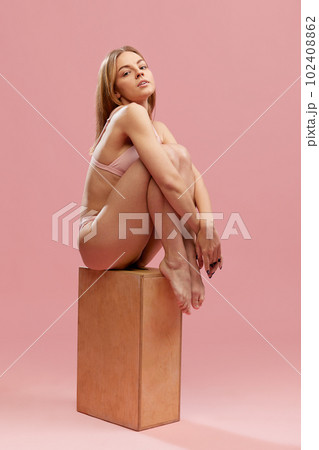 Portrait of young girl, woman wearing swimsuit sitting on wooden box and looking at camera with calm face over pink studio background. Self love 102408862