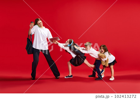 Portrait of businesswoman, mother talking on phone, little boy and girls, children pulling her hand, disturbing her against red studio background 102413272