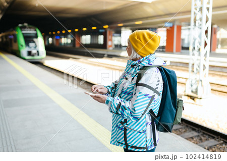 Old happy woman with backpack in yellow hat waiting train on station platform and using smart phone on urban background. Railroad transport concept Old happy woman with backpack in yellow hat waiting train on station platform and using smart phone on urban background. Railroad transport concept 102414819