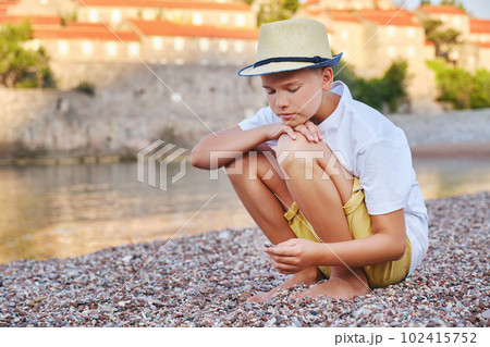 Sad or pensive stylish boy on the seashore examines pebbles. The concept of summer vacation and travel. 102415752