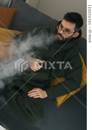 Bearded man is smoking hookah at home and blowing cloud of smoke - chill time and resting concept Bearded man is smoking hookah at home and blowing cloud of smoke - chill time and resting concept 102420031
