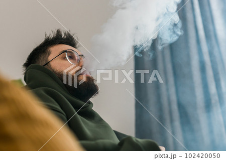 Bearded man is smoking hookah at home and blowing cloud of smoke - chill time and resting concept 102420050