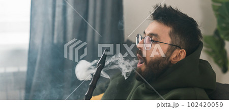 Banner bearded man is smoking hookah at home and blowing cloud of smoke copy space - chill time and resting concept 102420059