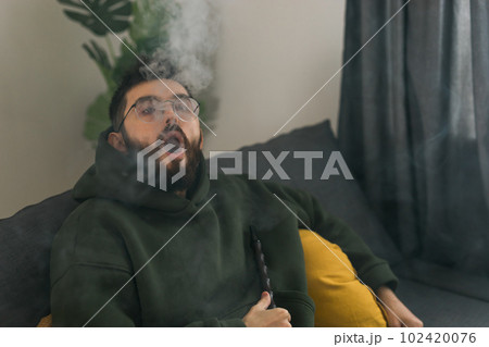 Bearded man is smoking hookah at home and blowing cloud of smoke - chill time and resting concept 102420076