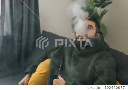Bearded millennial or gen z man smoking hookah while relaxing on sofa at home - chill time and resting concept Bearded millennial or gen z man smoking hookah while relaxing on sofa at home - chill time and resting concept 102420077