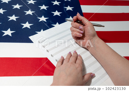 Female hands writing musical notes on a sheet of sheet music, against the background of the American flag, The concept of creating music Female hands writing musical notes on a sheet of sheet music, against the background of the American flag, The concept of creating music 102423130