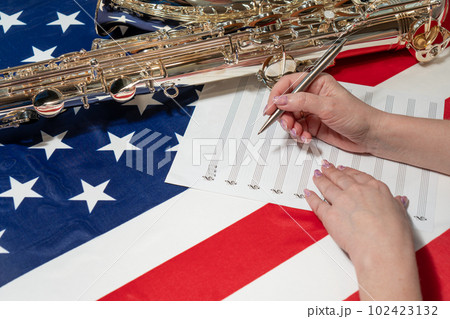 Female hands writing musical notes on a sheet of sheet music, against the background of the American flag, The concept of creating music Female hands writing musical notes on a sheet of sheet music, against the background of the American flag, The concept of creating music 102423132
