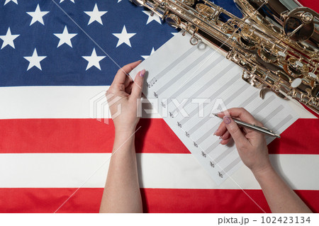 Female hands writing musical notes on a sheet of sheet music, against the background of the American flag, The concept of creating music Female hands writing musical notes on a sheet of sheet music, against the background of the American flag, The concept of creating music 102423134