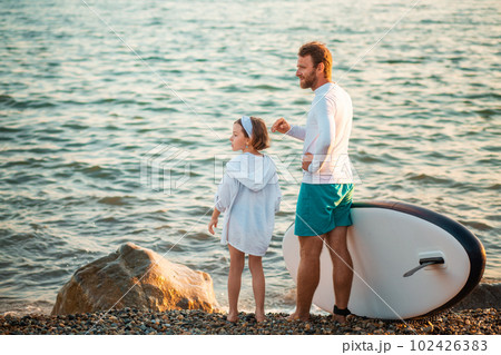Man and preschool girl standing on the beach with sup board. Ocean's surface on the background. Summer active sport at the sea. Back view Man and preschool girl standing on the beach with sup board. Ocean's surface on the background. Summer active sport at the sea. Back view 102426383
