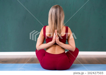 Blonde woman sitting in Lotus pose on blue mat putting hands into prayer gesture behind back lady in red sportswear enjoying yoga exercise and meditation in studio backside view 102427503