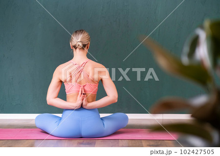 Female student practicing meditation for body and mind relaxation at professional yoga class young woman sitting in Lotus pose on pink mat with hands in prayer gesture behind back backside view 102427505