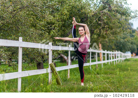 Full length portrait of flexible strong young girl stretching her leg, practicing yoga, having workout outdoor, raising leg in air, doing split stretch one foot, looking smiling away. 102429118