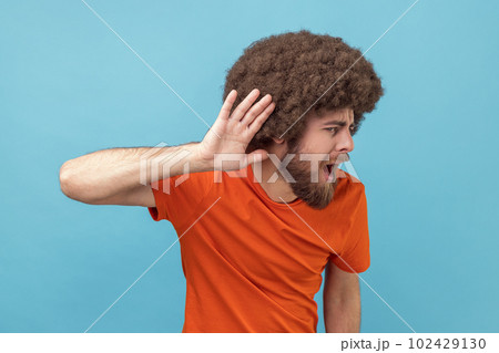 Portrait of man with Afro hairstyle in T-shirt holding hand near ear trying to listen quiet conversation, overhearing gossip, having hearing problems. Indoor studio shot isolated on blue background. Portrait of man with Afro hairstyle in T-shirt holding hand near ear trying to listen quiet conversation, overhearing gossip, having hearing problems. Indoor studio shot isolated on blue background. 102429130
