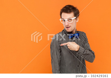 Portrait of man nerd pointing away on copy space, discusses amazing promo, gives way or direction, wearing shirt with blue bow tie and white glasses. Indoor studio shot isolated on orange background. 102429161