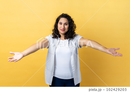 Please, take for free. Portrait of generous friendly woman with dark wavy hair welcoming with wide open arms and smiling kindly, happy to embrace you. Indoor studio shot isolated on yellow background. 102429203