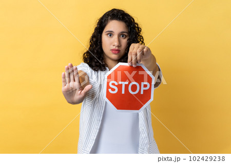 Portrait of strict woman with dark wavy hair holding red stop sign looking at camera with ban palm gesture, has strict expression, prohibition. Indoor studio shot isolated on yellow background. 102429238