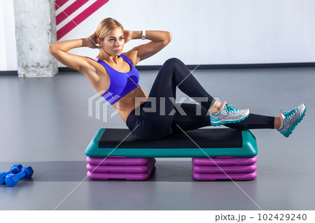 Side view portrait of sporty beautiful woman with blonde hair wearing tight sportswear sitting on step platform and doing press aerobic, having work out in fitness club. Indoor shot. 102429240