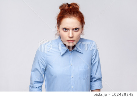 Portrait of angry aggressive ginger woman wearing blue shirt looking at camera with anger and hate, being in bad nervous mood. Indoor studio shot isolated on gray background. Portrait of angry aggressive ginger woman wearing blue shirt looking at camera with anger and hate, being in bad nervous mood. Indoor studio shot isolated on gray background. 102429248