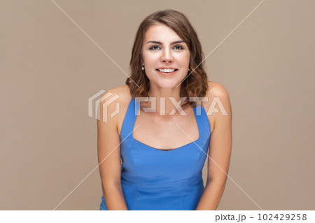 Portrait of smiling positive happy woman with wavy hair looking at camera with toothy smile, being in good mood, being glad, wearing blue dress. Indoor studio shot isolated on light brown background. Portrait of smiling positive happy woman with wavy hair looking at camera with toothy smile, being in good mood, being glad, wearing blue dress. Indoor studio shot isolated on light brown background. 102429258