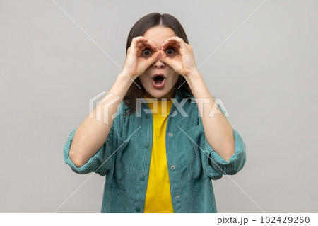 Shocked attractive young woman standing with hands on eyes binoculars gesture and looking at camera with surprised face, wearing casual style jacket. Indoor studio shot isolated on gray background. Shocked attractive young woman standing with hands on eyes binoculars gesture and looking at camera with surprised face, wearing casual style jacket. Indoor studio shot isolated on gray background. 102429260