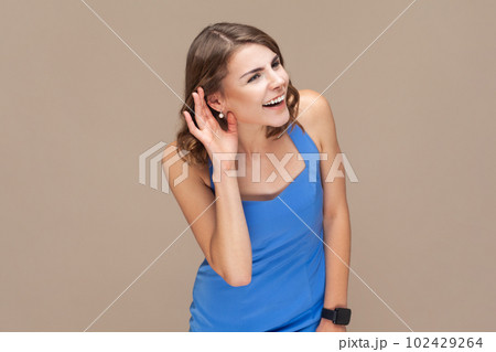 Portrait of attractive beautiful young adult woman with wavy hair standing palm near ear, listening private conversation, wearing blue dress. Indoor studio shot isolated on light brown background. 102429264