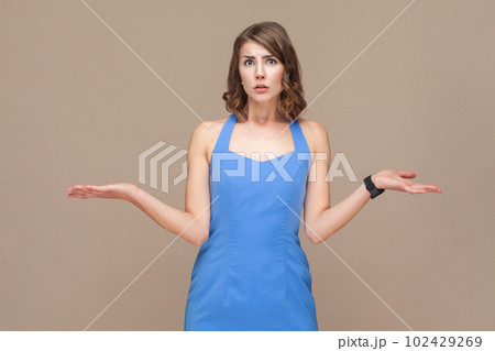 Portrait of puzzled confused young adult woman with wavy hair standing shrugging shoulders, don't know what to do, wearing blue dress. Indoor studio shot isolated on light brown background. 102429269