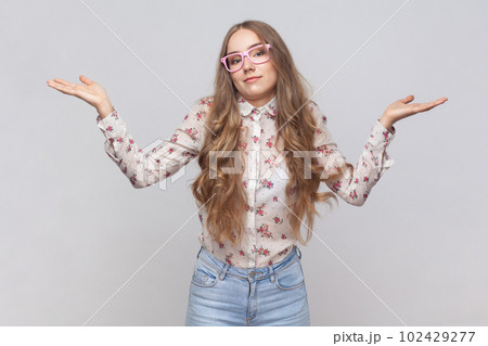 I don't know. Portrait of confused beautiful woman in glasses with wavy blond hair standing, raised arms, confused and thinking. Indoor studio shot isolated on gray background. 102429277