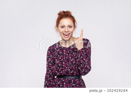 Portrait of astonished excited redhead woman wearing dress pointing finger up with genius idea, surprised by suddenly invented smart solution. Indoor studio shot isolated on gray background. 102429284