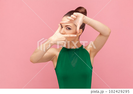 Portrait of concentrated woman photographer with bun hairstyle standing and looking at camera, making frame with fingers, wearing green dress. Indoor studio shot isolated on pink background. Portrait of concentrated woman photographer with bun hairstyle standing and looking at camera, making frame with fingers, wearing green dress. Indoor studio shot isolated on pink background. 102429295