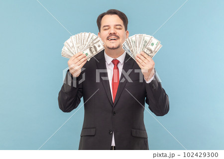 Portrait of greedy smiling man with mustache standing with closed eyes, holding dollar banknotes in hands, wearing black suit with red tie. Indoor studio shot isolated on light blue background. Portrait of greedy smiling man with mustache standing with closed eyes, holding dollar banknotes in hands, wearing black suit with red tie. Indoor studio shot isolated on light blue background. 102429300