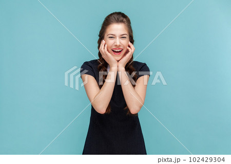 Portrait of funny joyful cheerful woman with braids standing with hands under chin and laughing, hearing joke, wearing black dress. woman Indoor studio shot isolated on blue background. 102429304