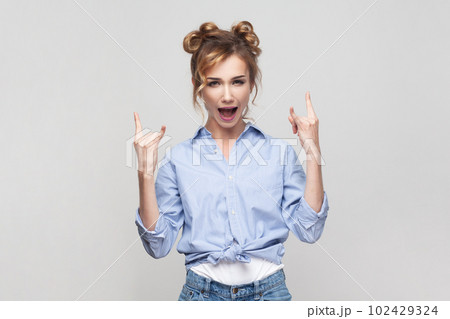 Portrait of blonde woman showing rock and roll gesture heavy metal sign enjoying favorite music on party has fun, exclaims from joy, wearing blue shirt. Indoor studio shot isolated on gray background. 102429324
