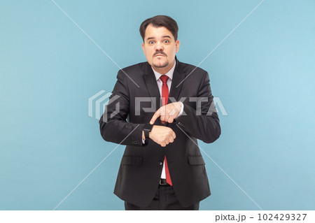 Portrait of serious man with mustache standing looking at camera, pointing at his smartwatch, deadline, wearing black suit with red tie. Indoor studio shot isolated on light blue background. 102429327