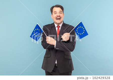 Man smiling broadly and holding flags of European Union, symbol of Europe, EU association and community, wearing black suit with red tie. Indoor studio shot isolated on light blue background. 102429328