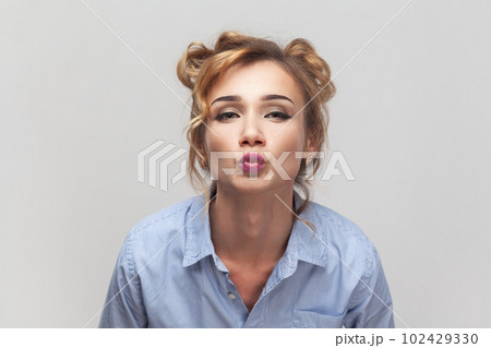 Air kiss for you. Portrait of blonde woman keeps lips folded, expressing love, blowing mwah, having romantic expression, wearing blue shirt. Indoor studio shot isolated on gray background. 102429330