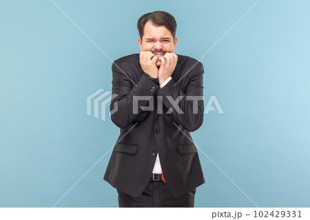 Portrait of nervous worried man with mustache standing biting his fingernails, having serious problems, scared, wearing black suit with red tie. Indoor studio shot isolated on light blue background. 102429331
