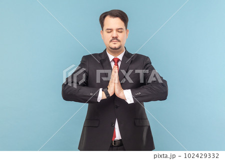 Portrait of calm man with mustache standing with closed practicing yoga, meditating while having break at work, wearing black suit with red tie. Indoor studio shot isolated on light blue background. 102429332