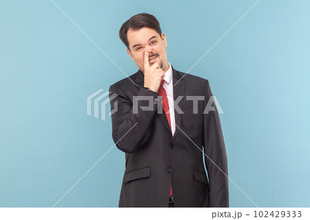 Portrait of funny childish man with mustache standing having comical facial expression, keeps finger in nose, wearing black suit with red tie. Indoor studio shot isolated on light blue background. 102429333