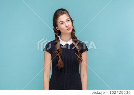 Portrait of pensive thoughtful woman with braids standing and looking away, making decision, thinks about solving problem, wearing black dress. woman Indoor studio shot isolated on blue background. 102429336