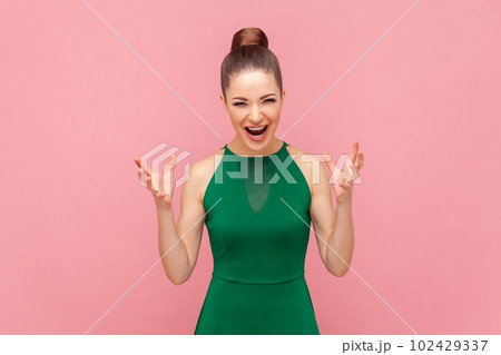 Portrait of crazy angry woman standing with raised arms, screaming with aggression, having problems, shouting, wearing green dress. Indoor studio shot isolated on pink background. 102429337
