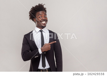 Man with Afro hairstyle has happy expression, smiles gently, points aside with fore finger, shows place to hang out, wearing white shirt and tuxedo. Indoor studio shot isolated on gray background. 102429338