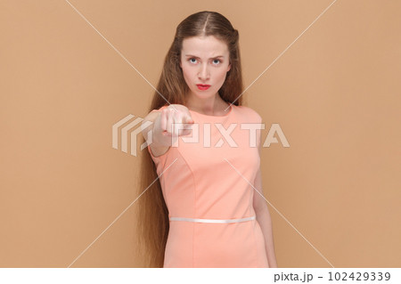 Portrait of serious strict woman with long hair standing pointing to camera, choosing you, select, making choice, wearing elegant dress. Indoor studio shot isolated on brown background. 102429339