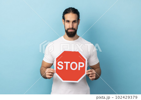 Portrait of serious man with beard wearing white T-shirt holding red Stop sign, looking at camera with negative aggressive expression, showing ban. Indoor studio shot isolated on blue background. 102429379