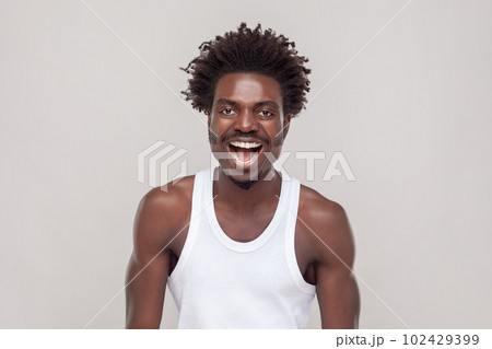 Emotional gorgeous man with Afro hairstyle grinning broadly, enjoying good day, having fun, laughing at something funny, wearing white T-shirt. Indoor studio shot isolated on gray background. 102429399